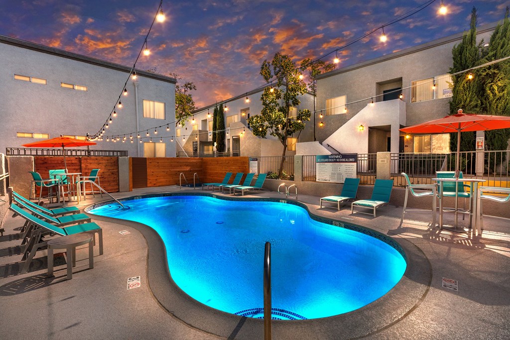 Colton, CA Apartments - Ardella At Reche Canyon - Dusk View Of Swimming Pool With Lounge Seats, Tables, Chairs, Umbrellas, And Cafe Lights.