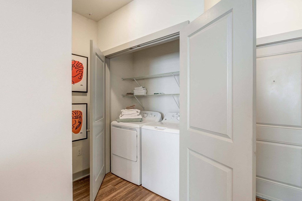 Apartment laundry closet with stacked washer and dryer, open white doors, and shelves with folded towels.