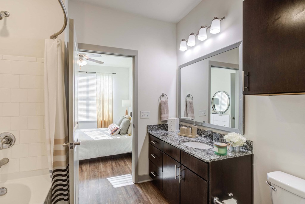 Apartment bathroom with granite countertop, dark wood cabinets, and view into bedroom.