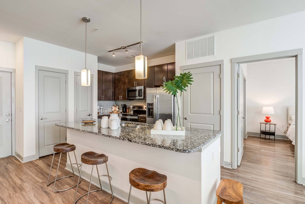 Apartment kitchen with granite countertop island, wooden stools, stainless steel appliances, and view into bedroom.