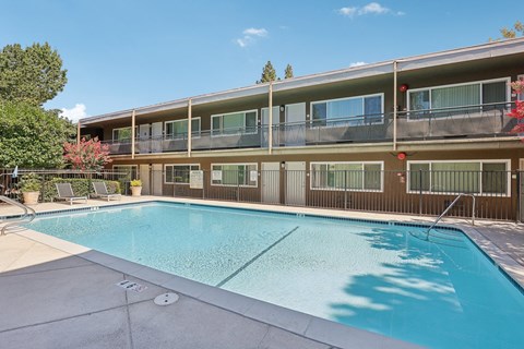 Townhomes in Walnut Creek, CA - Carmel House Apartments - Swimming Pool With Clear Water, Lounge Chairs, Metal Fencing, Surrounding Apartment Building, Balconies, Trees, And Blue Sky.
