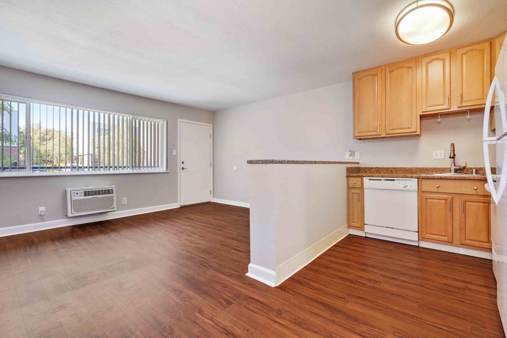 Downtown Walnut Creek CA Apartments - Carmel House Apartments - Living Room Area with Open-Concept Kitchen Layout, Wood Inspired Floors, Wall Heater/AC, Window with Blinds, and Door