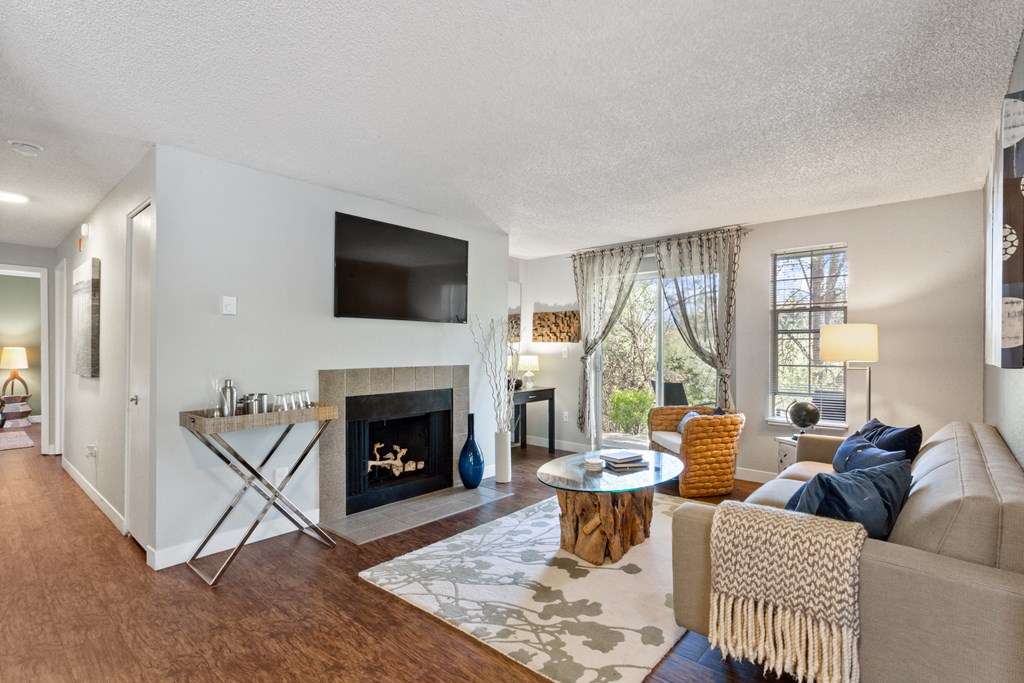 2-Bedroom Apartments in Beaverton, OR - Cedar Crest - Living Room with Vaulted Ceilings, a Fireplace, Wood-Inspired Flooring, Mounted TV, Cozy Couch, and a Coffee Table with Decorative Area Rug and Access to Patio.