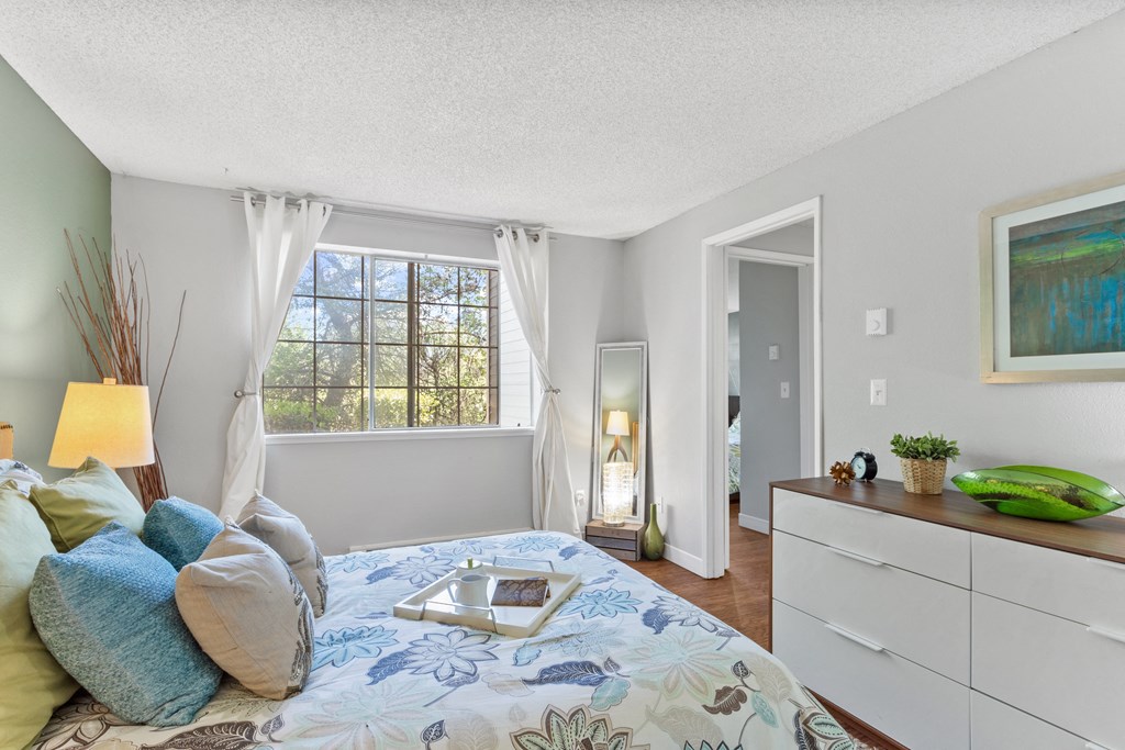 Apartments For Rent in Beaverton, OR - Cedar Crest - Bedroom with Large Window, Bed with White Comforter with Blue Flowers, Dresser, and Wood-Inspired Flooring.