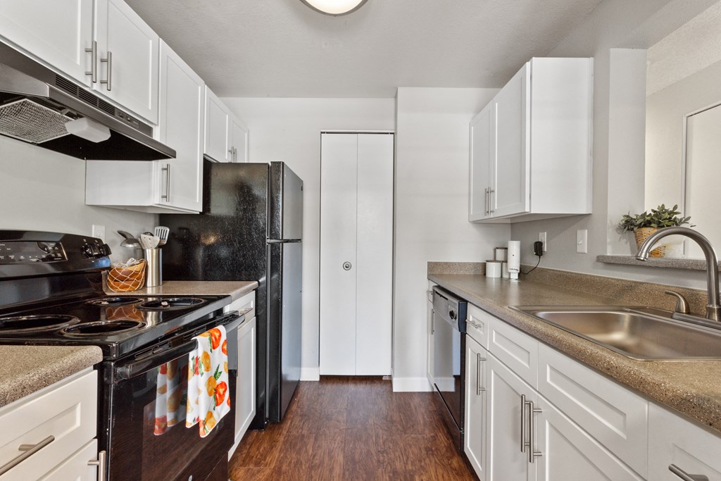 Apartments in Beaverton, OR, For Rent - Cedar Crest - Kitchen with Wood-Inspired Flooring, White Cabinets, Large Sink, Dishwasher, and Black Appliances.