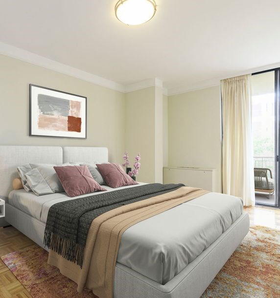 A bed with a white headboard and a grey blanket is in the foreground of a room with a framed picture on the wall and a chair by the window.