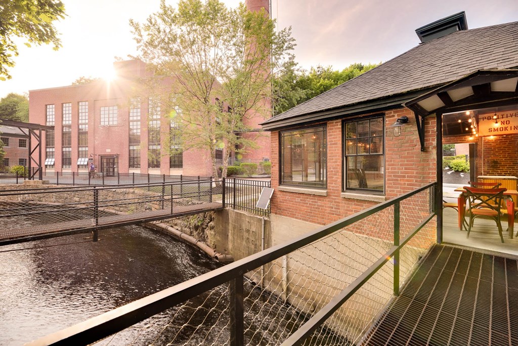 Brick pavilion with outdoor seating overlooking a river and connected to a brick building by a metal bridge.