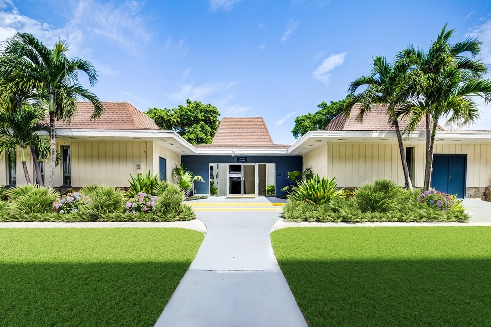 Entrance to the leasing office surrounded by palm trees and landscaped lawns at Coronado Springs Apartments in Palm Springs, FL. Pet-friendly community with one- and two-bedroom apartments.