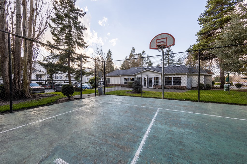 Apartments In Kent, WA, With In-Unit Washer And Dryer - Country Glen - A Fenced Basketball Court Surrounded By Trees With Nearby Houses And A Trash Can Along The Side.