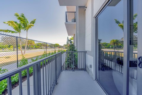 Apartment balcony with metal railing overlooking a street with palm trees and a chain-link fence.