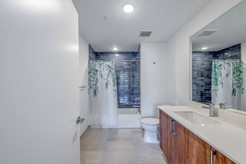 Modern bathroom with blue tiled shower, leaf-patterned curtain, white toilet, and wood-grain vanity.