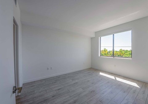 Empty, bright apartment bedroom with light gray wood-look flooring and a window view of palm trees.