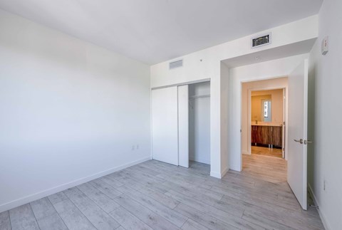 Empty apartment bedroom with white walls, wood-look flooring, closet, and bathroom view.