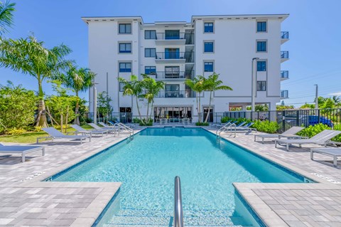 Apartment building with a swimming pool, lounge chairs, and palm trees on a sunny day.
