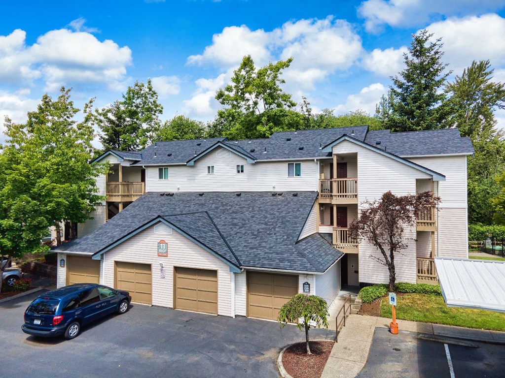 University Place, WA, Garden-Style Apartments with In-Unit Washer and Dryer - Carriage House - An Exterior View of the Attached Garages, an Open Parking Area, and a View of the Apartments, Landscaping, and Trees.