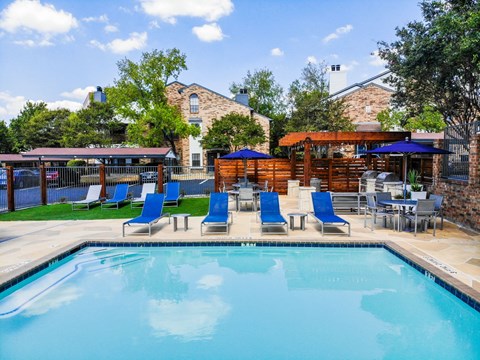 Austin, TX, Townhomes, With In-Unit Washer and Dryer - Cottages at Wells Branch - Resort Style Pool With Blue Lounge Chairs, Shade Umbrellas, Patio Seating, and Grilling Station Under a Pergola.