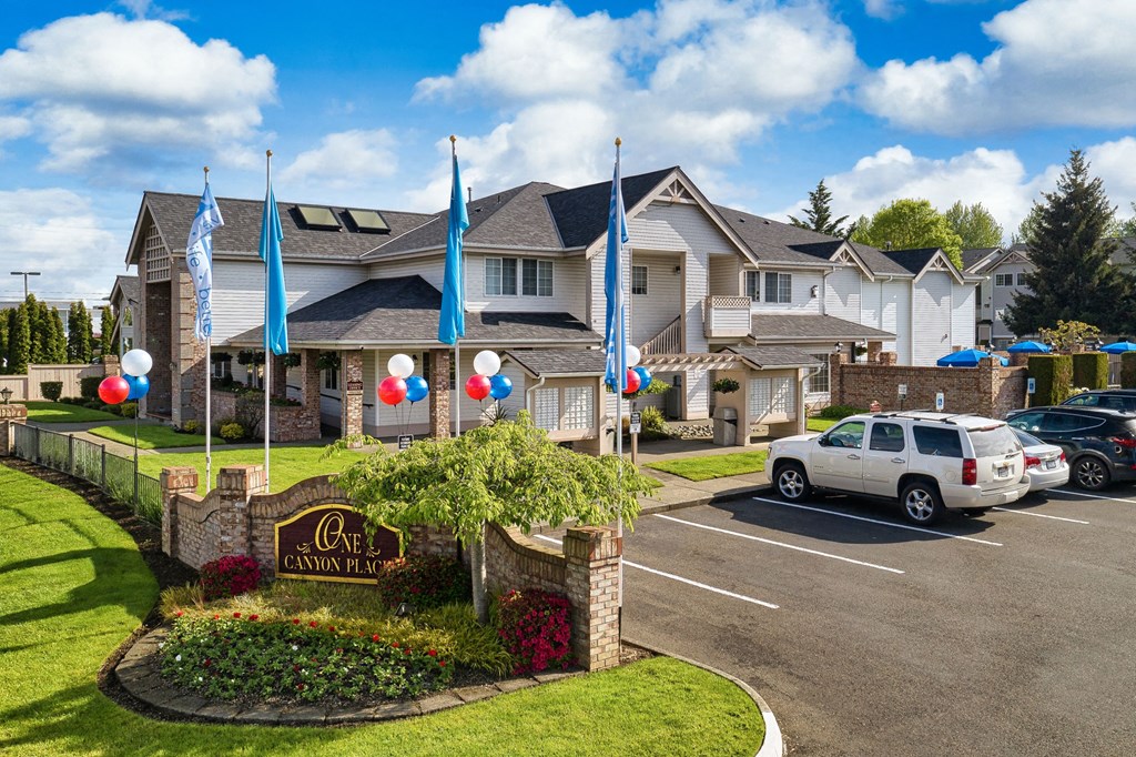 Apartments for Rent in Puyallup, WA - One Canyon Place - Exterior View of Property with Signage, Flags, Balloons, Manicured Lawn, Parking Lot, and View of Apartment Community.