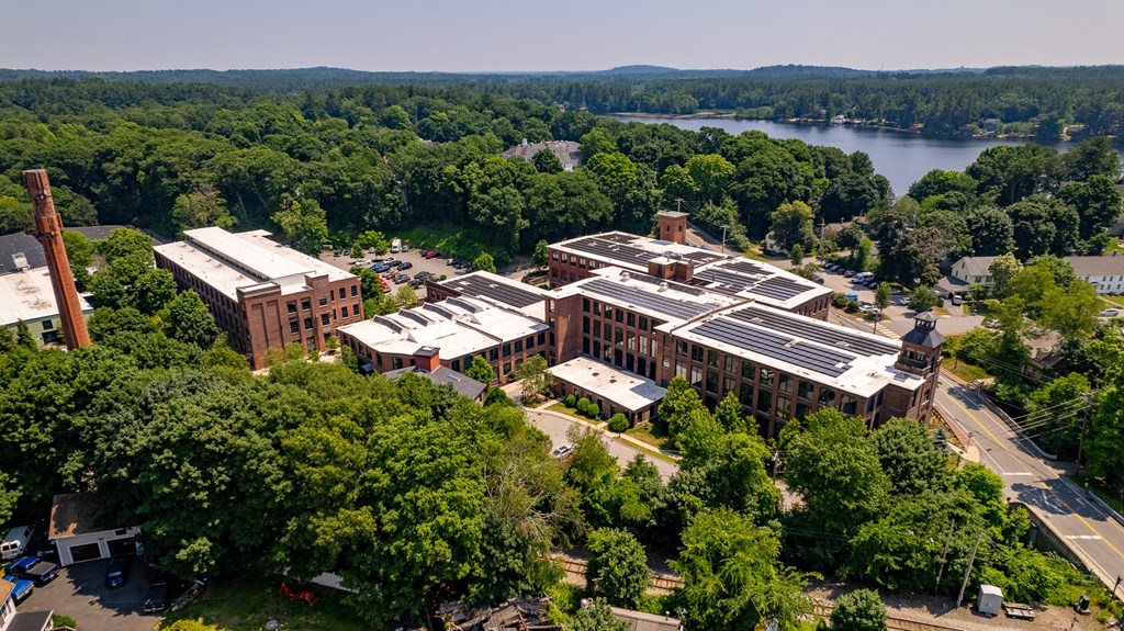 Aerial view of a brick mill complex with solar panels, surrounded by lush green trees and a lake in the background.