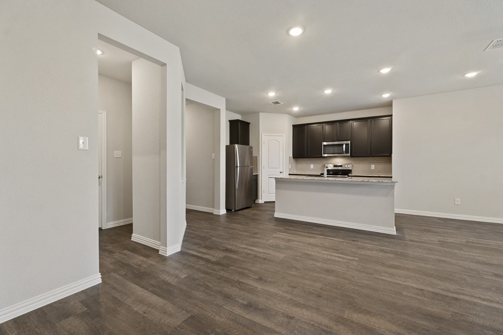 an empty living room with a kitchen with a refrigerator and a sink