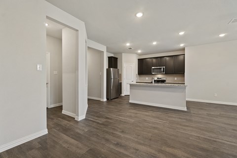 an empty living room with a kitchen with a refrigerator and a sink