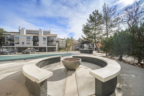 Apartments in Aurora, CO, with an In-Unit Washer and Dryer - The Juliet at Lowry - A Fire Pit Surrounded by a Circular Concrete Bench, Metal Fence, Trees, and a View of the Parked Cars and Apartments.