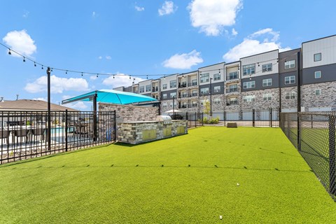 A green artificial turf soccer field with a fence and a building in the background.