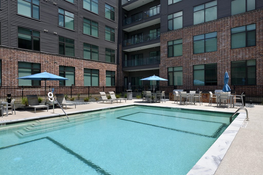 A swimming pool surrounded by chairs and umbrellas in front of a brick building.