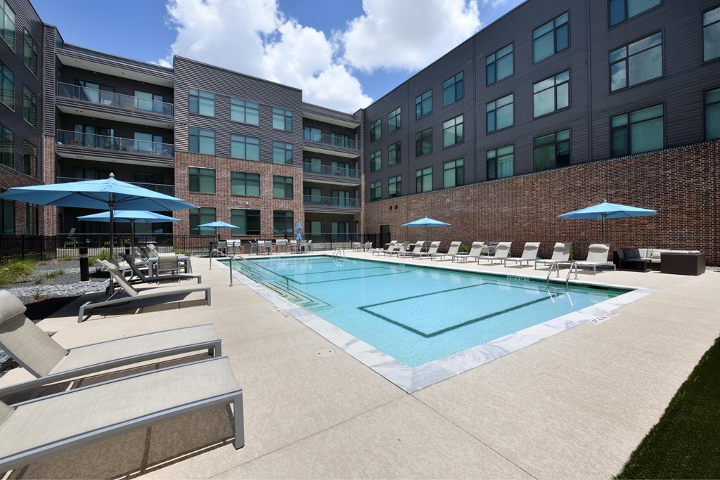 A swimming pool surrounded by sun loungers and umbrellas in front of a brick building.