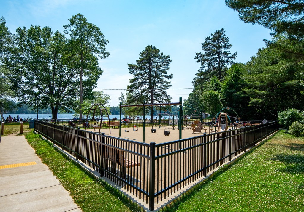 Fenced playground with swings, slides, and climbing structures near a lake.