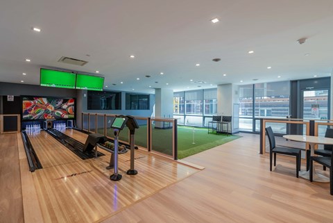 The bowling alley amenity space at Encore 325 Apartments features glossy wood lanes, a green turf area for games, and large windows with natural light.