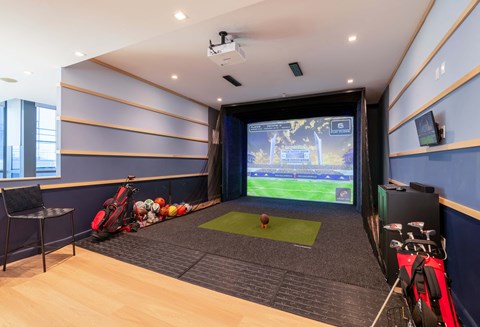 The indoor golf simulator room at Encore 325 Apartments features a projector screen displaying a green course, a striking mat, a golf bag, and blue padded walls.