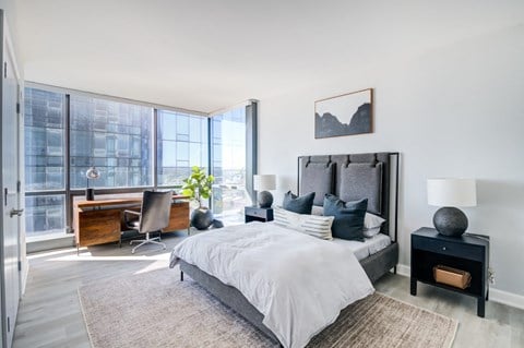 A luxurious and bright bedroom features a gray upholstered bed, a large corner window with a city view, and an abstract black and white painting above the headboard.