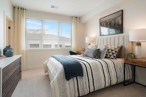 Reunion, CO, Garden-Style Apartments Near Barr Lake State Park - Enova - Bedroom With Upholstered Bed, Neutral Carpet, Bedside Tables, Lamps, Wall Art, and Large Window With Coverings.