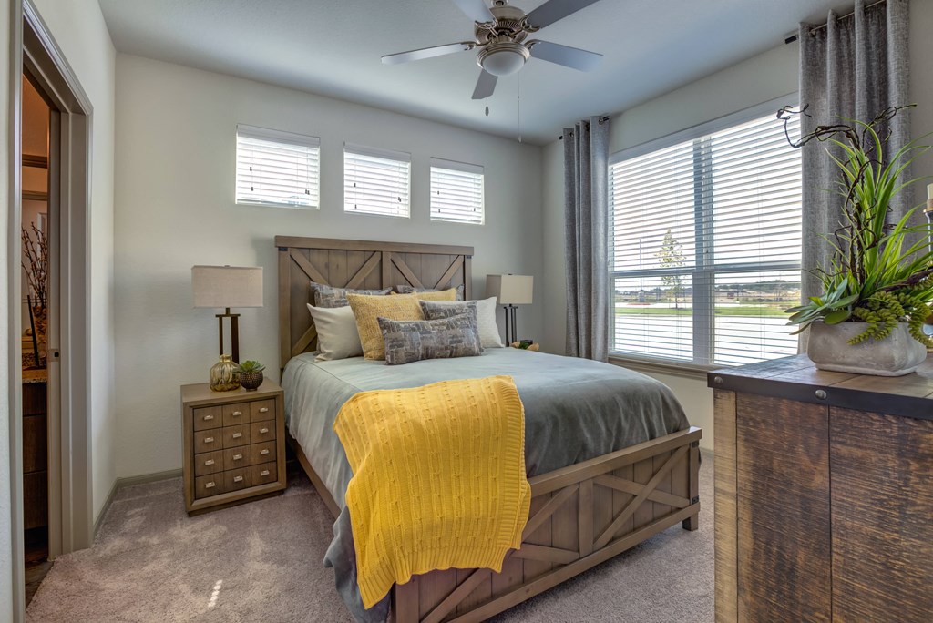 One Bedroom Apartments in Georgetown Texas - Williamson at the Overlook - Furnished Bedroom with Bed, Wood-Style Frame, Nightstand, and Dresser, with Window, Ceiling Fan, Carpetted Floors, and Attached Bathroom View