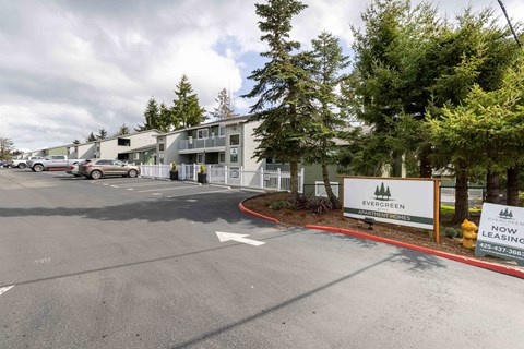 Lynnwood, WA, Apartments - Evergreen Grove - Parking Lot with Vehicles, Trees, Fence, Apartment Buildings, and Property Signage.