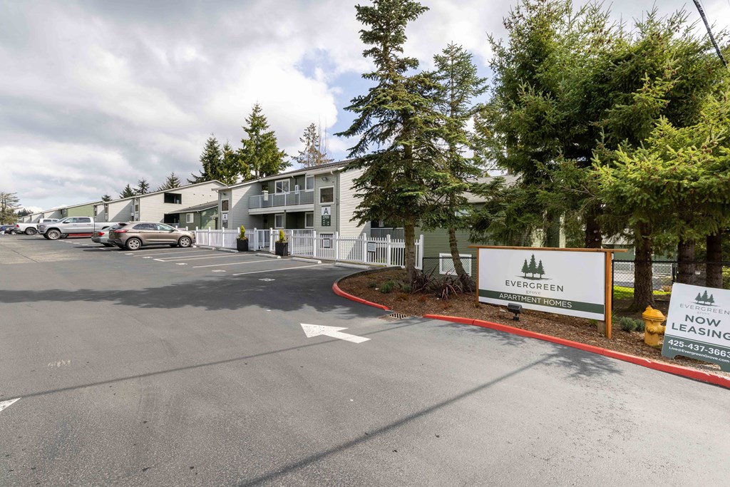 Lynnwood, WA, Apartments - Evergreen Grove - Parking Lot with Vehicles, Trees, Fence, Apartment Buildings, and Property Signage.