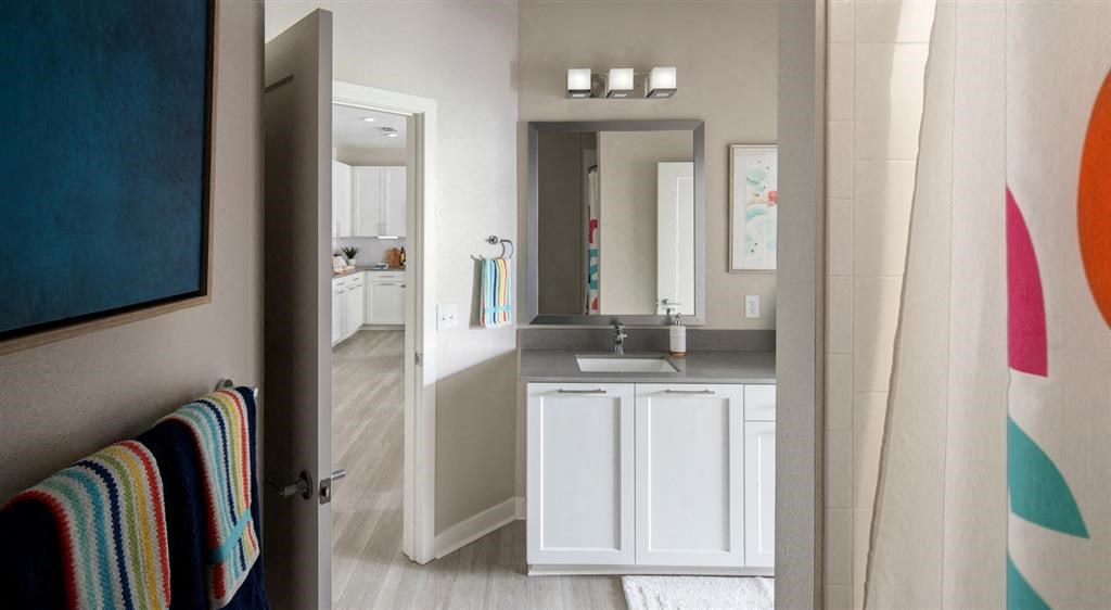 Modern bathroom with white vanity, gray countertop, colorful shower curtain, and view into kitchen.