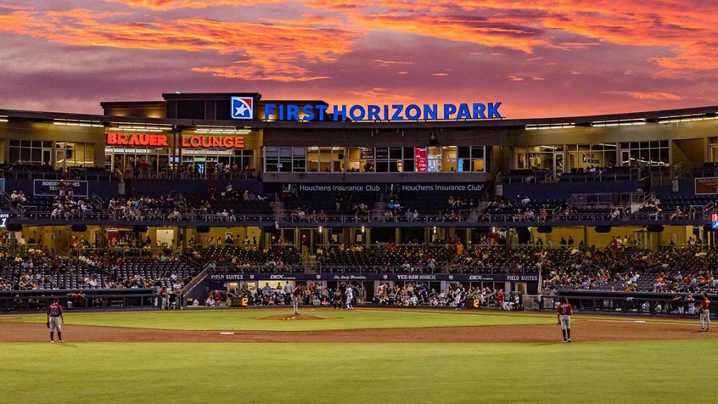 Baseball game in progress at a stadium with a colorful sunset sky.