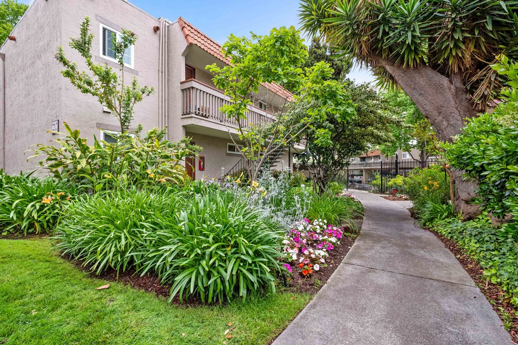 Two Bedroom Apartments in Hayward CA - Glen Oaks - Concrete Path Surrounded by Grassy Lawns and Landscaped Flowers, Trees, and Hedges, with Tan Building Exterior Nearby