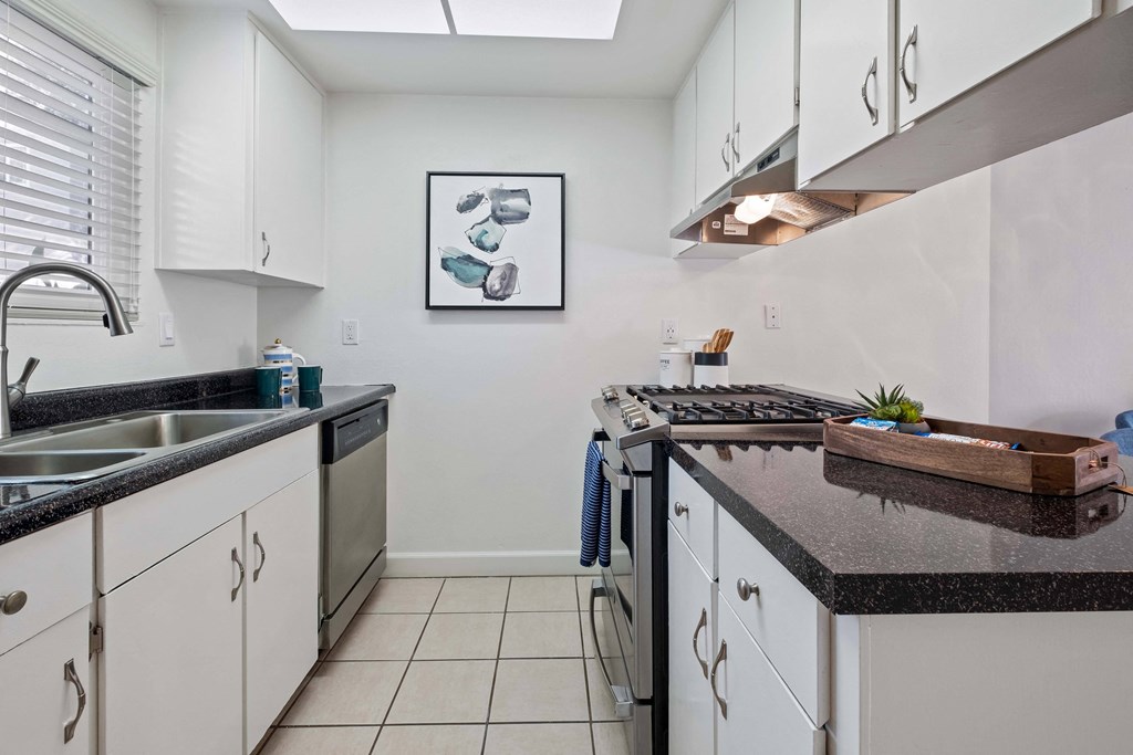 Hayward, CA Apartments for Rent - Glen Oaks - Modern Aisle-Style Kitchen with Black Granite Countertops, Stainless Steel Appliances, Tiled Flooring, White Cabinetry, and a Nearby Window