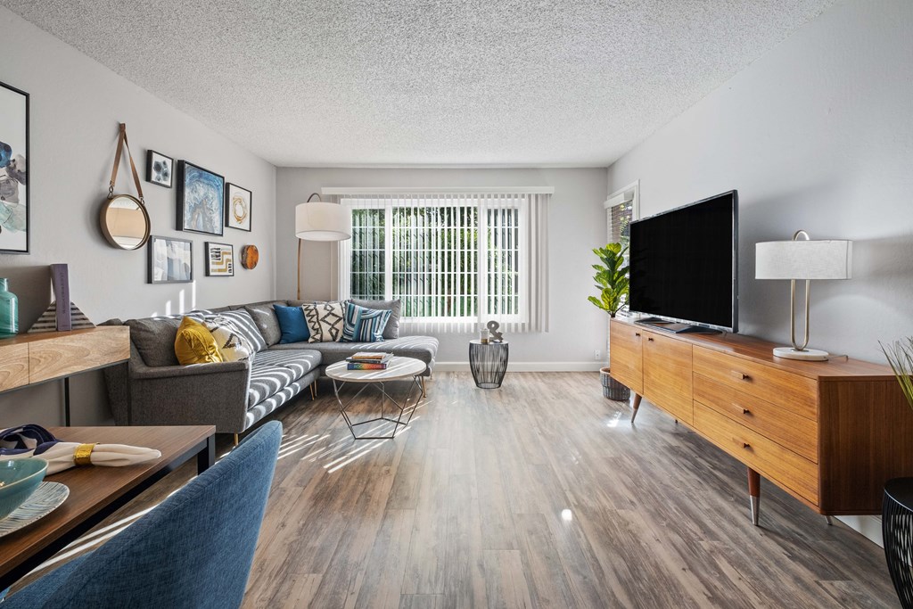 Dog-Friendly Apartments in Hayward CA - Glen Oaks - Furnished Living Room with Grey L-Shaped Couch, Wood-Style Flooring, Round Coffee Table, Wooden Desk and Chair, Moutned Shelving, Wooden Storage Cab