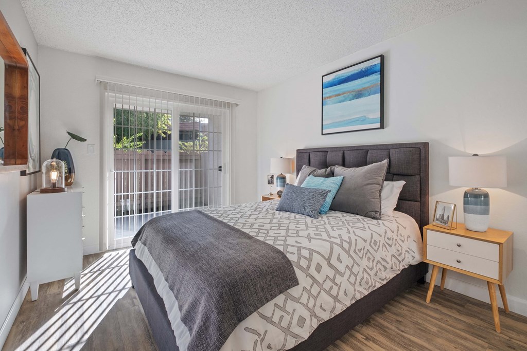 One Bedroom Apartments in Hayward CA - Glen Oaks - Spacious Bedroom with Wood-Style Flooring, a Bed, Wooden Nightstands with Lamps, White Dresser, and Large Window/Sliding Door with Patio Access