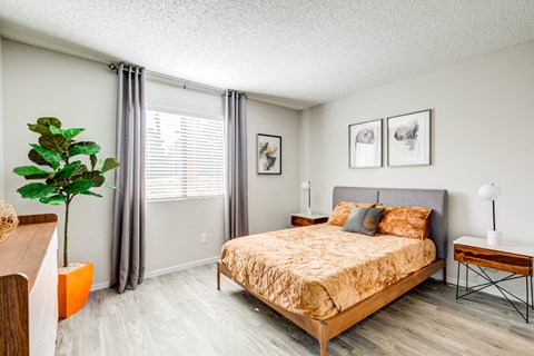 Senior Living Las Vegas, NV - Gramercy Parc Senior Apartments - Bedroom With Gray Headboard, Wood-Style Flooring, Large Window, Gray Curtains, Nightstands, And Neutral Walls.