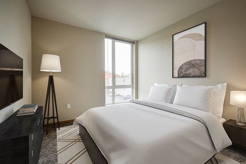 One-Bedroom Apartments in South Seattle, WA - Greenhouse - Bedroom with Carpeted Flooring, Bed, Wooden Nightstand with Lamp, Standing Lamp, Wooden Dresser, Mounted TV, and Extra-Large Window.