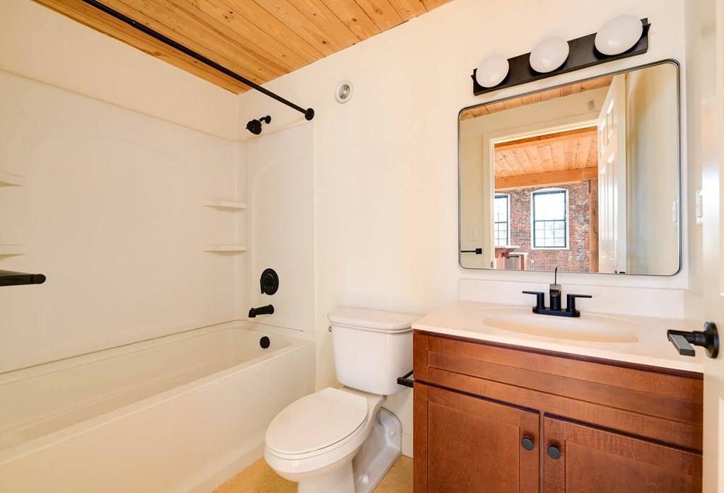 Bathroom with white tub and shower, toilet, and wood vanity with sink, reflecting a room with exposed brick and wood ceiling.