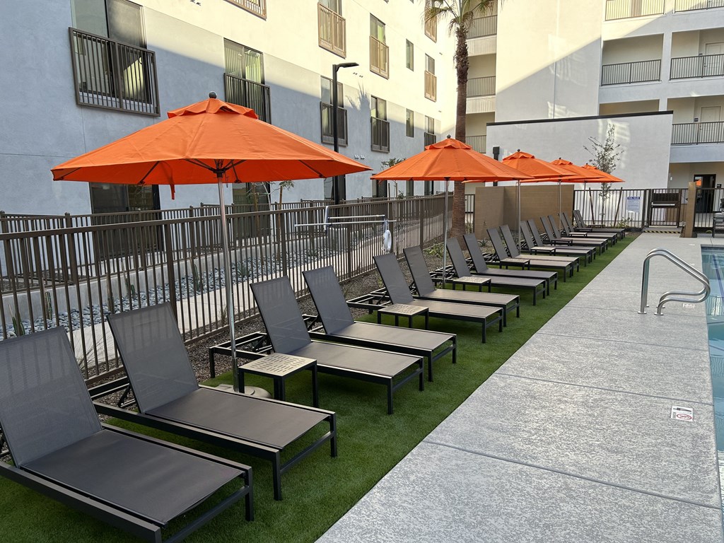 Studio Apartments in Downtown Phoenix, AZ - VB on 12th Street - Poolside Lounge Seating with Orange Umbrellas on a Patch of Artificial Grass Turf and Surrounded by a Fence.