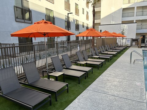 Downtown Phoenix, AZ, Apartments Near ASU - VB on 12th Street - Poolside Lounge Area With Orange Umbrellas, Chaise Seating, Turf Lawn, And Apartment Building Views.