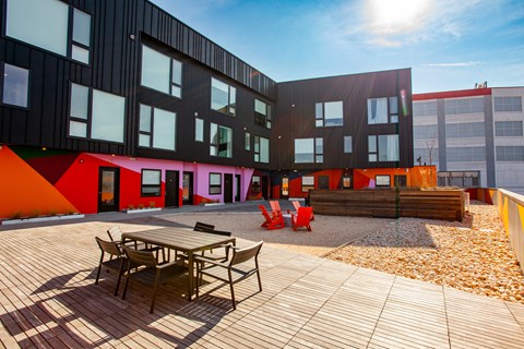 A wooden table and chairs are on a patio in front of a building with black and red walls.