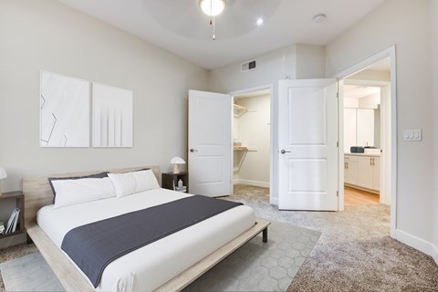 1-Bedroom Apartments in Milpitas, CA - Ilara Apartments - Bedroom With White Bedding, Dark Throw Blanket, Open Closet, Ceiling Fan, Nightstands, Wall Art, Carpeted Flooring, And Bathroom Access.