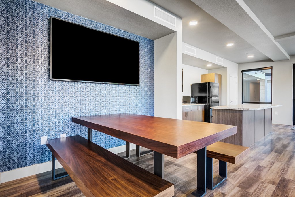 Studio Apartments in Phoenix, AZ - Imperial - Community Space with Wood-Style Flooring, Picnic-Style Table and Benches, Wall-Mounted TV, Kitchen, and Window.
